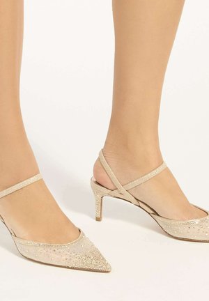 Beige Slingback-Pumps mit spitz zulaufender Zehenpartie, dünnen Riemchen und funkelnden Strassverzierungen, getragen an den Füßen vor schlichtem Hintergrund.