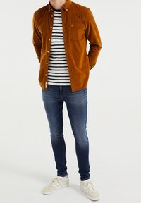 Brun kordflöjelskjorta med knappar över en svartvit randig t-shirt, parat med blå jeans och beige sneakers.