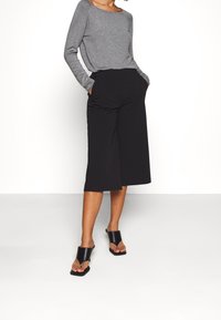 Maglia grigia a maniche lunghe con scollatura rotonda, abbinata a culotte nere a gamba larga. Sandali con tacco aperti neri completano l'outfit.