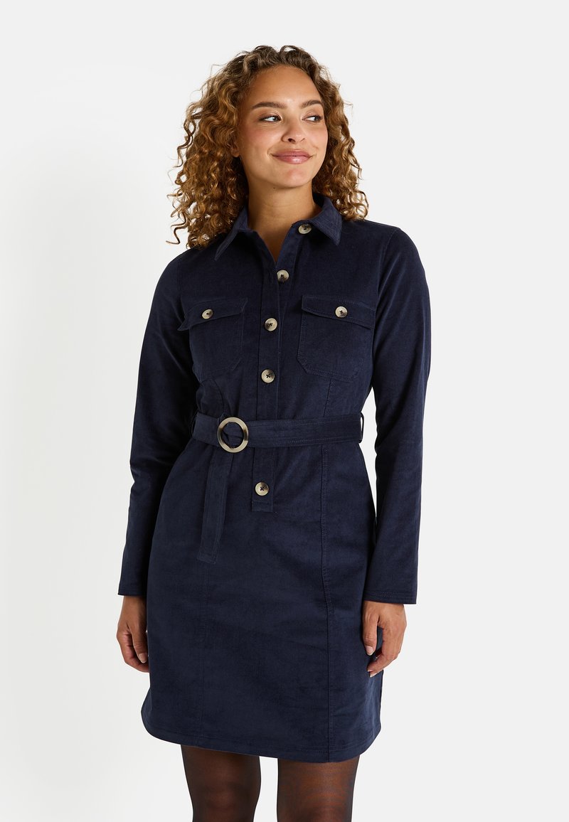 Femme aux cheveux bouclés portant une robe longue à manches longues, ceinturée, de couleur bleu marine, avec des détails de boutons et deux poches poitrine, se tenant devant un fond blanc.