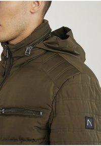 Oliven grøn pufferjakke med høj krave, lynlås-lommer, teksturerede skuldre og logo-patch på ærmet. Strømlinet design, let materiale.