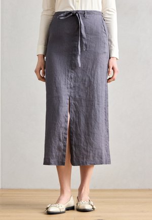 Jupe en lin gris avec fente à l'avant et détail de taille nouée, associée à des chaussures plates de couleur claire. Le tissu semble texturé et ample.