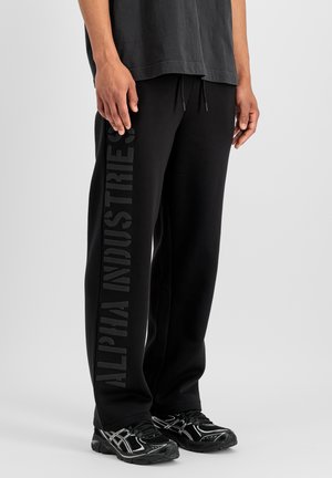 Uomo che indossa pantaloni della tuta neri con "ALPHA INDUSTRIES" stampato verticalmente sulla gamba sinistra e scarpe da ginnastica nere con dettagli bianchi.