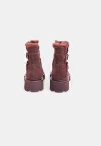 Botas de tobillo de ante marrón con un acabado texturizado, que cuentan con un cuello esponjoso, dos hebillas y una suela gruesa y estriada.