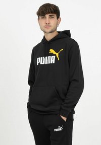Sudadera negra con bolsillo canguro, que presenta un gran logotipo blanco de "PUMA" y una gráfica de un puma amarilla saltando en la parte frontal. Material de algodón.