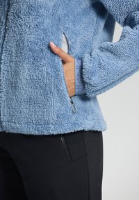 Person, der eine hellblaue, flauschige Jacke trägt, mit der Hand in der Seitentasche, kombiniert mit schwarzen Hosen, die eine Reißverschlusstasche haben.