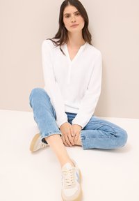 Hvid langærmet skjorte med V-hals og knaplukning, parret med lyseblå jeans med opkrøllede ben og beige sneakers med blå accenter.