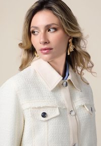 Giacca bianca con texture e colletto, pattina frontale beige a contrasto e due tasche sul petto. Presenta bottoni argentati e un tessuto liscio.
