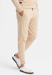 Beige Jogginghose mit seitlichen Taschen, schmalem Schnitt, gerippten Bündchen und kontrastierenden schwarzen Details. Kombiniert mit weißen Sneakers.
