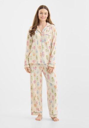 Vrouw die een crèmekleurige pyjama draagt met meerkleurige hondenprints, een top met knopen en een lange broek, staand op blote voeten tegen een witte achtergrond.