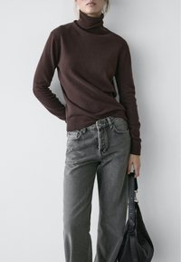 Bruine coltrui van zachte gebreide stof, gecombineerd met loszittende grijze spijkerbroeken. Zwarte handtas in één hand gehouden.