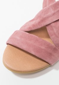 Sandale plate à bout ouvert avec une semelle marron clair et de larges brides en daim rose doux croisées sur le pied.