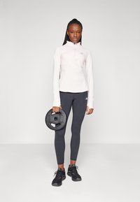 Maglietta rosa chiaro a maniche lunghe con mezzi zip, leggings neri con logo, disco pesante nero e scarpe da ginnastica nere. Texture liscia, design aderente.
