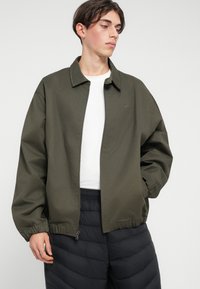 Aliejinės žalios spalvos audinio švarkas su apykakle ir užtrauktuku. Savybė – elastinės rankovės ir subtilus logotipas ant krūtinės. Dėvimas ant balto marškinio.