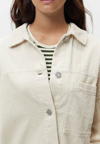 Cremefarvet kordfløjl jakke med knaplukning, en brystlomme og ribbet tekstur, båret over en grøn og hvid stribet bluse.