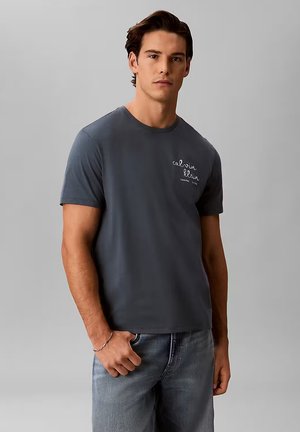 Giovane uomo che indossa una t-shirt Calvin Klein grigio scuro e jeans blu, in piedi davanti a uno sfondo grigio chiaro uniforme.