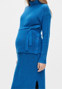Pull en maille côtelée bleue et jupe assortie, avec un col roulé, une ceinture nouée et des boutons décoratifs sur le côté du pull.