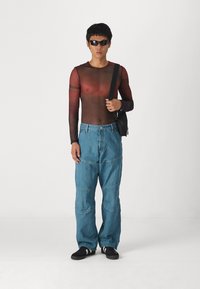 Top comprido justo e transparente em gradação de vermelho a castanho, combinado com jeans de denim azul claro, folgados, com detalhes em patchwork. Óculos de sol pretos e uma mala.