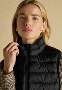 Fekete puffervest magas gallérral és cipzárral, vízszintes varrással. Egy kockás ing fölött viselt, gombokkal.