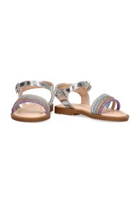 Sandalias metálicas plateadas con suelas planas. Presentan tiras de rhinestone multicolores en púrpura, azul y transparente, con hebilla ajustable en el tobillo.
