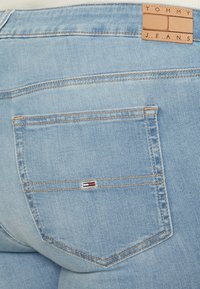 Helle blaue Denim-Jeans mit einer einzelnen Gesäßtasche, versehen mit orangefarbenen Nähten und einem kleinen gestickten Logo in Rot, Weiß und Blau.