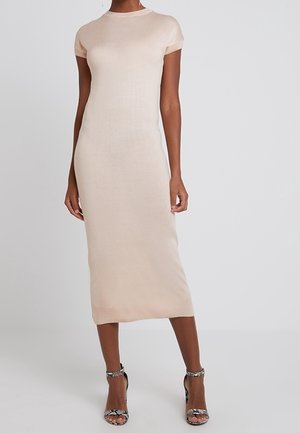 Kobieta w dopasowanej, beżowej sukience midi z krótkimi rękawami, w połączeniu z sandałami na wysokim obcasie i otwartymi palcami o wzorze wężowej skóry, stojąca na gładkim tle.