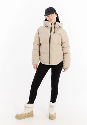 Beige pufferjack met een hoge kraag en zwarte ritsen, gecombineerd met zwarte leggings en crèmekleurige winterlaarzen met bontafwerking.