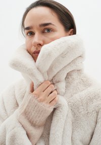 Faux fur-jacka i mjuk gräddfärg med hög krage, med en plyschtextur och synliga sömmar, lagd över en ljusrosa stickad tröja.