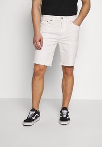 Bílé džíny šortky s roztrhaným lemem, mají pět kapes a kovové detaily. Vybavené černými teniskami a černým tričkem.