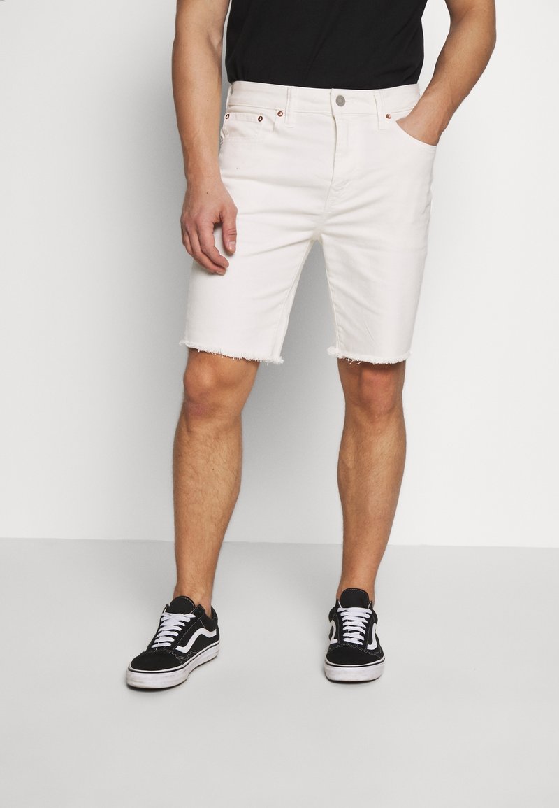 Bílé džíny šortky s roztrhaným lemem, mají pět kapes a kovové detaily. Vybavené černými teniskami a černým tričkem.