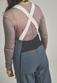 Blue pants with black waist and white straps featuring "ORTOVOX" text; purple long-sleeve shirt with a subtle gradient texture. Zipper detail visible.