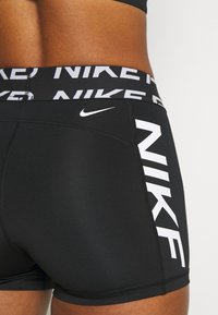 Černé sportovní šortky s širokým elastickým pasem s opakujícím se nápisem "NIKE" a bílým logem Nike Swoosh na boku.