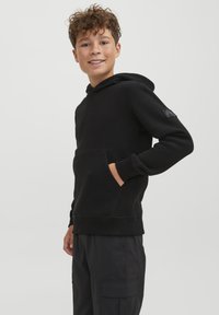 Felpa nera con cappuccio realizzata in tessuto morbido, con tasca frontale e polsini a costine, indossata con pantaloni scuri.