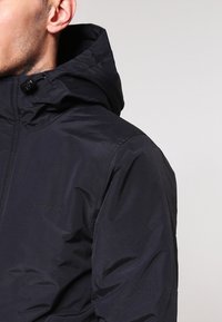 Sort hættejakke med en glat tekstur, der har lynlås lukning, justerbar kant og diskret branding på forsiden.