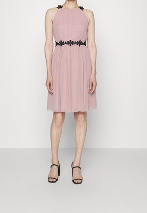 Femme portant une robe rose clair sans manches avec des détails floraux noirs sur les bretelles et à la taille, associée à des sandales noires à talons.