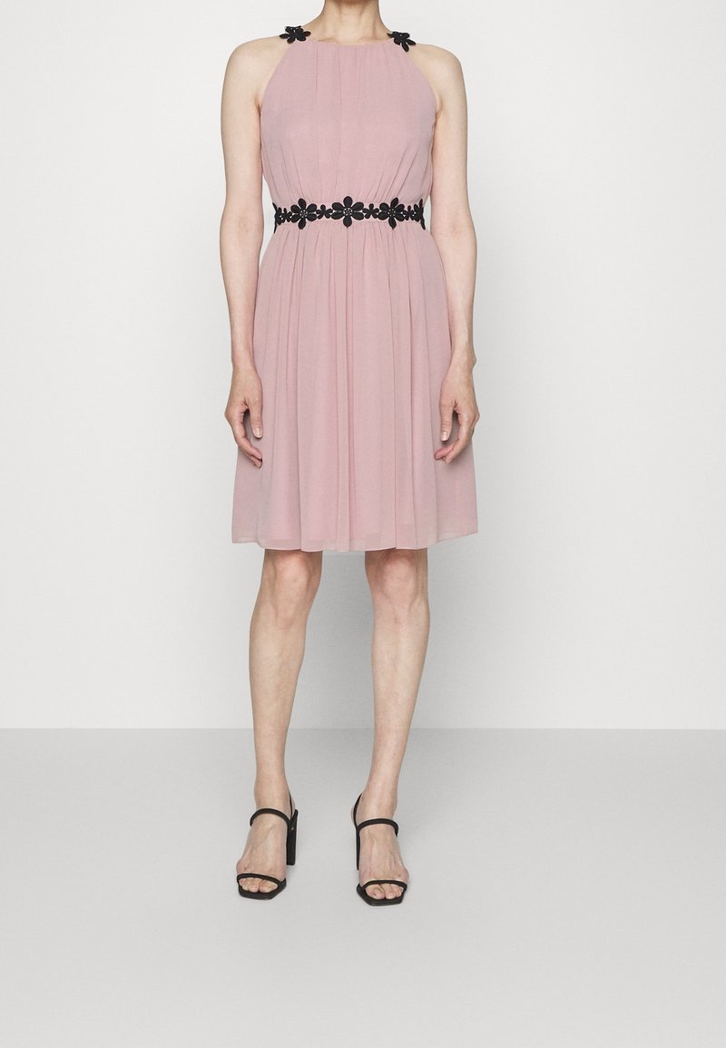 Femme portant une robe rose clair sans manches avec des détails floraux noirs sur les bretelles et à la taille, associée à des sandales noires à talons.