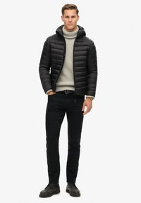 Chaqueta tipo puffer negra con capucha, diseño acolchado, llevada sobre un suéter de cuello alto gris claro, combinada con jeans negros y zapatos oscuros.