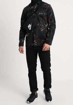 Chemise à boutons à motif camouflage dans des tons de vert foncé, rouge et gris ; associée à un jean noir et des baskets noires avec accents blancs ; sac bandoulière.
