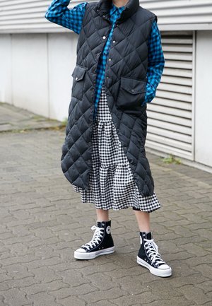Zwarte gestikte lange vest over een blauw geruit shirt, gecombineerd met een zwart-wit geblokte rok en zwarte hoge sneakers.
