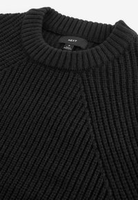Pull en tricot noir avec une texture côtelée, montrant le col et l'étiquette portant la mention "NEXT", fabriqué au Bangladesh, taille Medium.