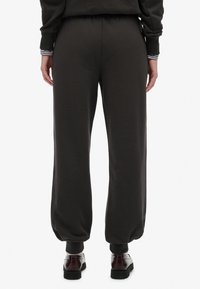 Persoon in zwarte losvallende joggingbroek en donkerbruine schoenen, staand met ontspannen handen langs hun zij, gezien vanaf de achterkant.