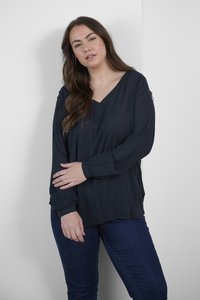 Mörkblå blus med V-ringning, långa ärmar och subtil textur. Matchad med mörkblå jeans, står mot en ljus bakgrund.