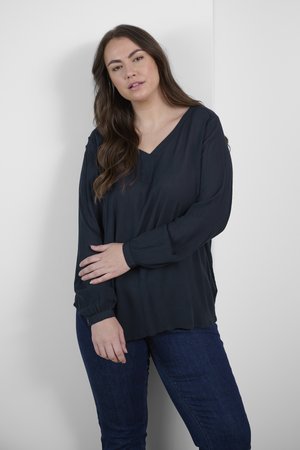 Blusa blu scuro con scollatura a V, maniche lunghe e una texture sottile. Abbinata a jeans blu scuro, in piedi contro uno sfondo chiaro.
