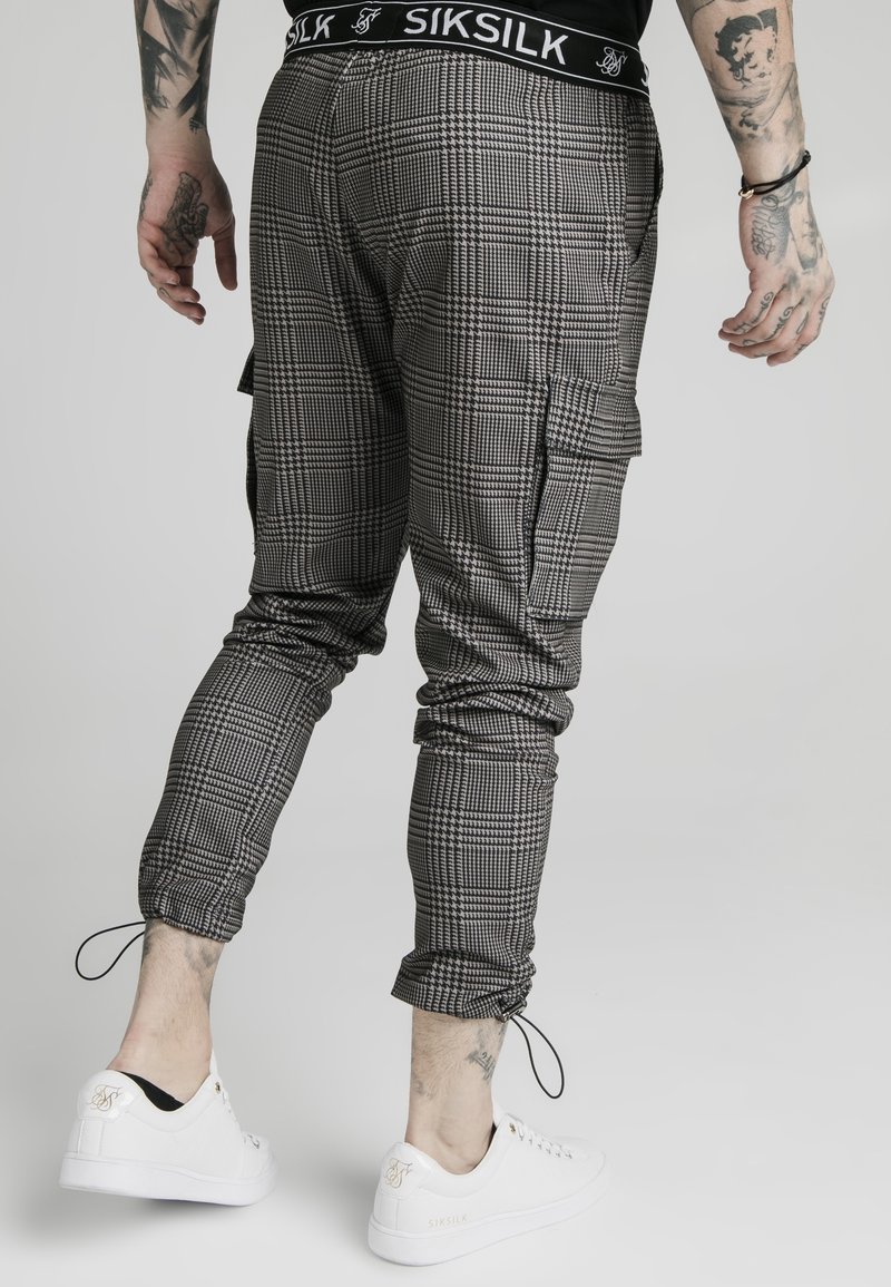Black and white checkered cargo pants with tapered fit, drawstring ankles, and a black waistband featuring "SIKSILK" logo. White sneakers visible.