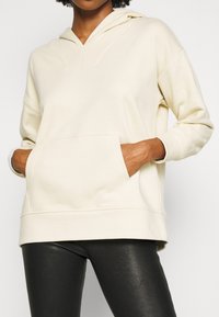 Felpa color crema con design a V, tasca frontale e maniche lunghe. Tessuto morbido con una vestibilità comoda, abbinata a leggings neri aderenti.