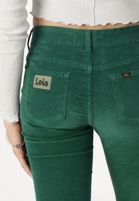 Green corduroy pants with a fitted design. Features a branded patch on the back pocket. Textured fabric with vertical ribbing and visible seams.