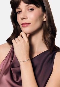 Srebrna bransoletka z akcentami w kształcie gwiazdek z przezroczystymi kamieniami szlachetnymi. Noszona na dłoni, uzupełniona satynową purpurową bluzką.