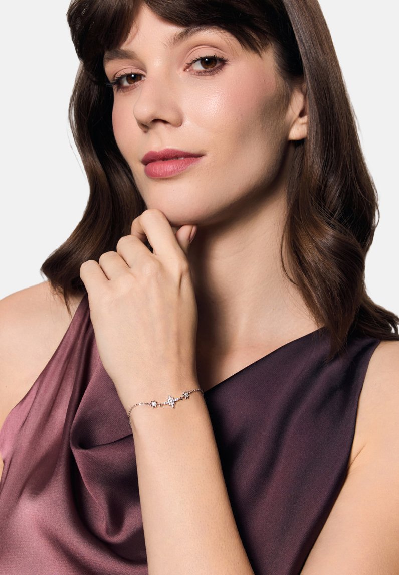 Srebrna bransoletka z akcentami w kształcie gwiazdek z przezroczystymi kamieniami szlachetnymi. Noszona na dłoni, uzupełniona satynową purpurową bluzką.