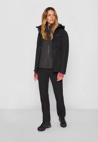 Ženska oblečena v črno zunanjo jakno, temno sivo zadrgo flis, črne hlače in črne pohodne čevlje stoji ob enotni ozadju.