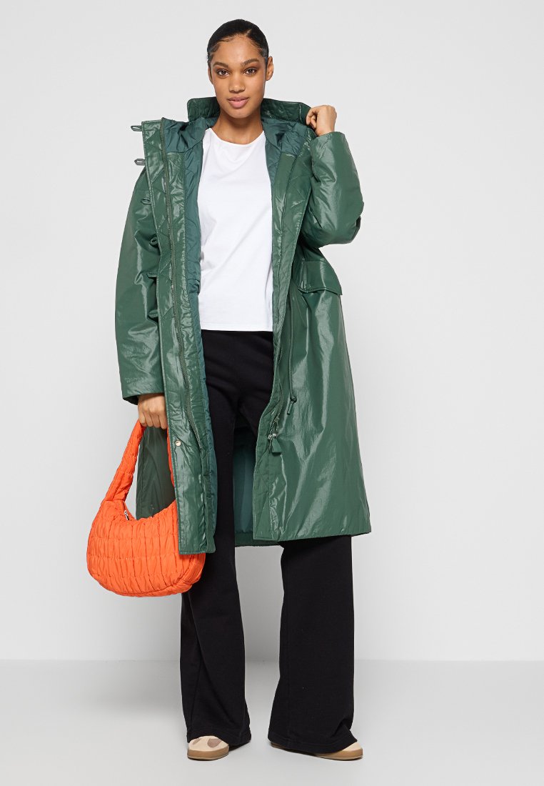 Parka impermeable verde con cremallera, cordones y dos bolsillos frontales, combinada con pantalones negros y un bolso acolchado naranja.